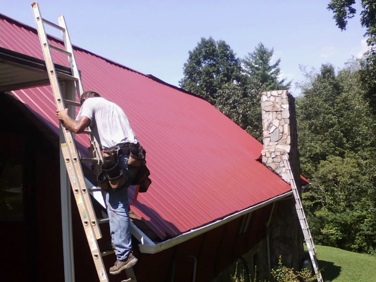 Roofer on a ladder beside a red metal roof and stone chimney — Soffit & Fascia Repair by Roofing Brothers in Horsham